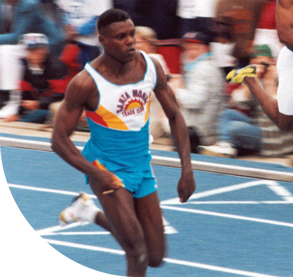 Close-up action shot of a male runner in a blue and white Santa Monica Track Club singlet sprinting on a blue track during a competition.
