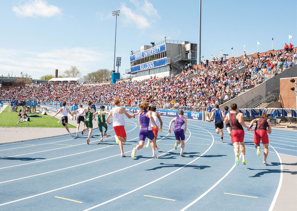 Low-angle view from behind relay runners as they sprint around a blue track curve toward the Drake University Bulldogs press box and packed bleachers.
