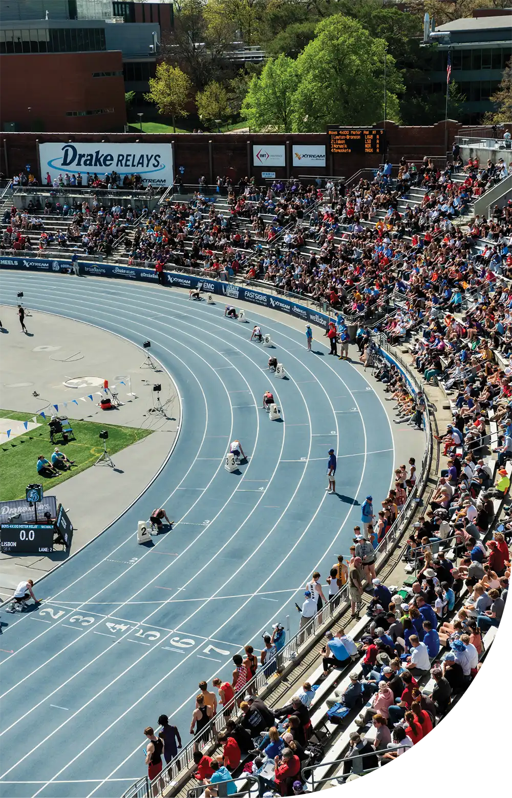 High-angle shot of athletes in starting blocks on a bright blue track curve during the Drake Relays, with spectators filling the stands in the background.