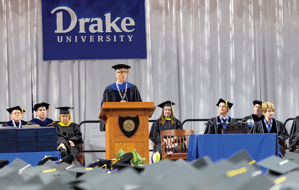 A speaker in academic regalia addresses a crowd from a wooden lectern at a Drake University ceremony. Faculty sit behind him against a white backdrop with a blue university sign.