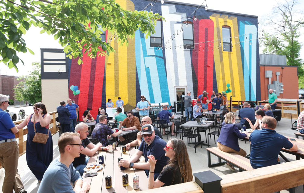 People gather at outdoor patio tables and benches in front of a building featuring a large, colorful "DOGTOWN" mural. The atmosphere is casual with string lights and blue and green balloons.