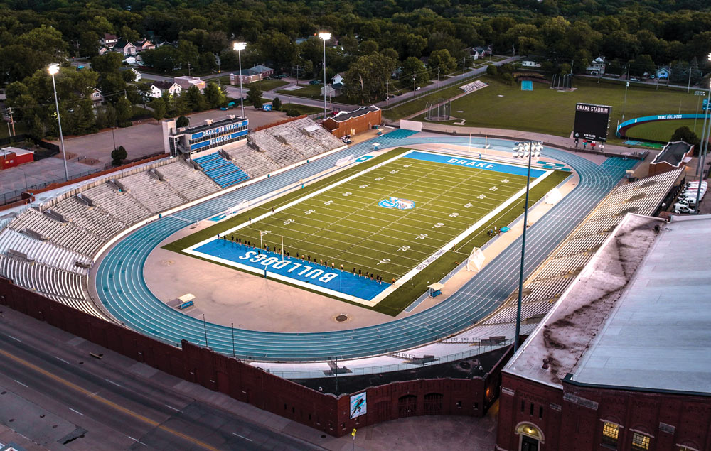 An elevated twilight view of Drake Stadium, showing the bright green football field, blue end zones, and the surrounding blue running track under glowing stadium lights.
