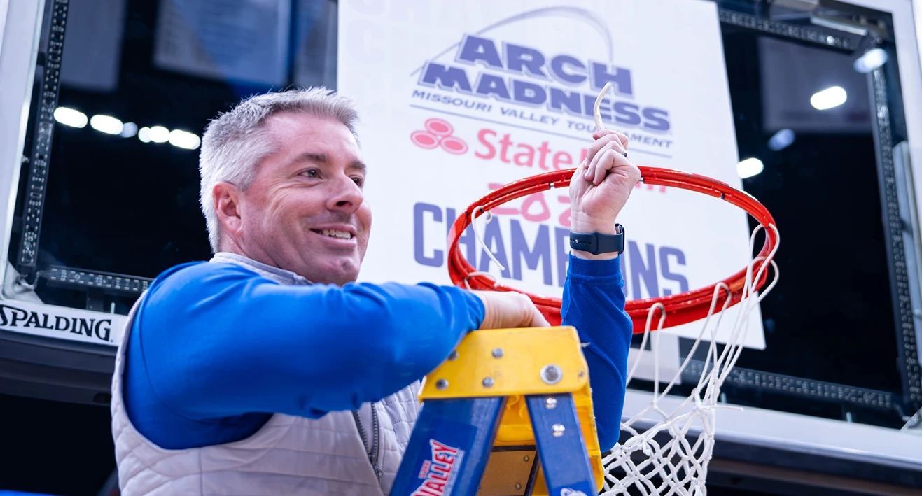 Man standing on a ladder cutting down a basketball net after a championship game, smiling as he holds the net. A sign behind him reads “ARCH MADNESS Missouri Valley Tournament” and “State Farm,” with the word “CHAMPIONS” visible below, marking a tournament victory celebration.