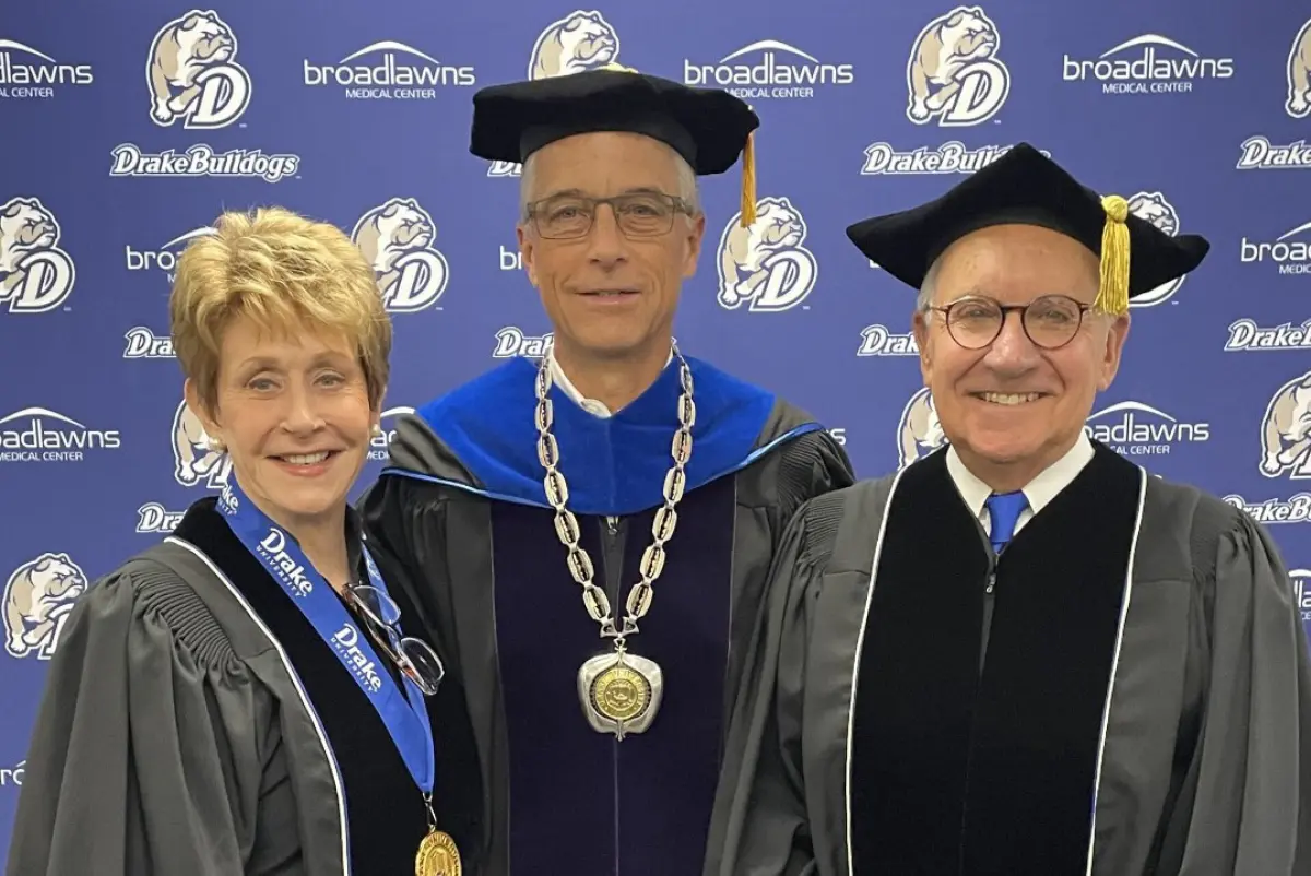 A formal photo of three people in academic regalia standing in front of a blue backdrop with Drake University Bulldogs and Broadlawns Medical Center logos; The woman on the left and the man on the right wear black robes, while the man in the center wears a dark purple and blue robe with a large silver ceremonial chain