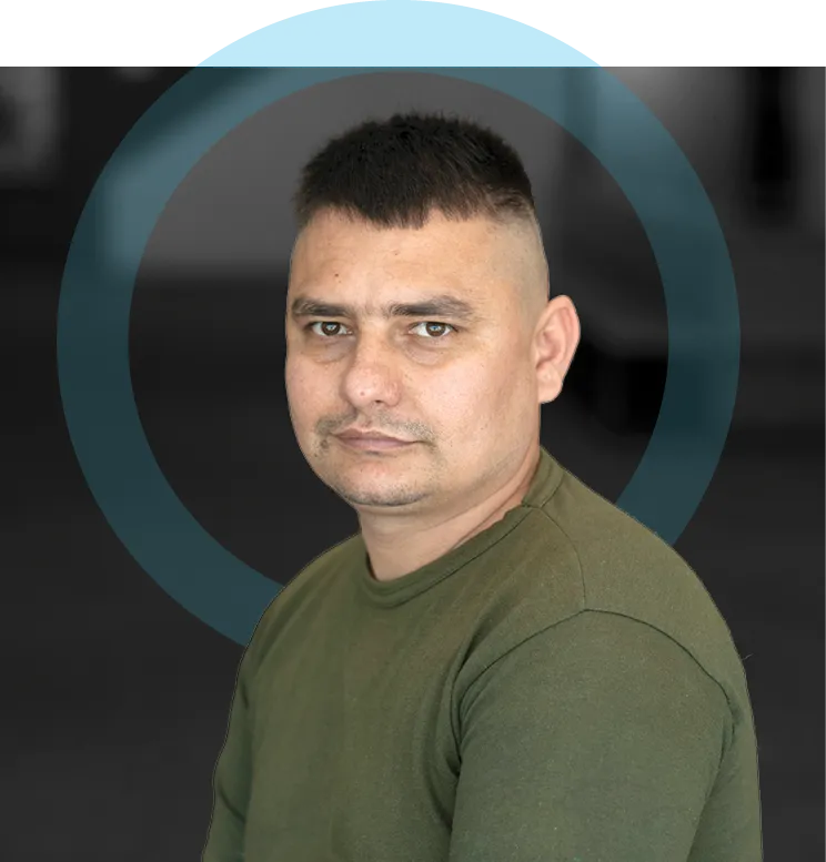 Studio portrait of an adult man with short dark hair wearing a green long-sleeve shirt, seated and facing the camera with a neutral expression against a dark background with a circular graphic element.