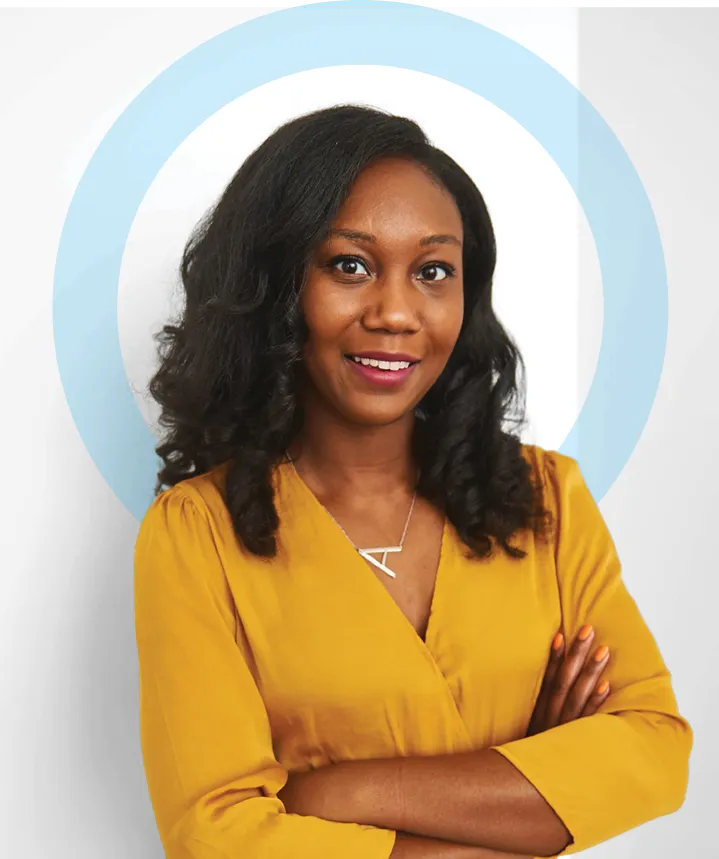 Studio portrait of an adult woman with shoulder-length dark hair wearing a mustard yellow blouse, arms crossed and smiling slightly, posed against a light background with a circular graphic element.
