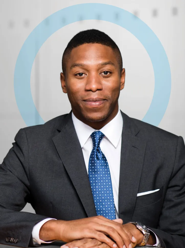 Studio portrait of an adult man wearing a dark suit, white shirt, and blue tie, seated with hands folded and looking directly at the camera against a light background with a circular graphic element.