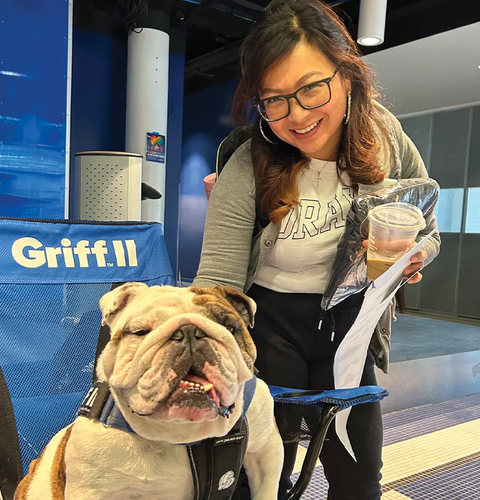 A woman wearing glasses smiles while standing beside Griff II, Drake University’s bulldog mascot, who is seated in a chair indoors; she holds a drink and papers during a campus event.