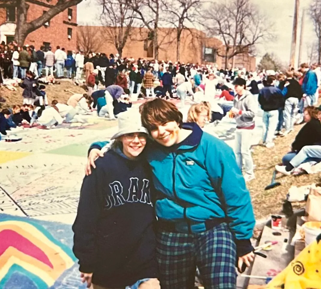 Two students stand with their arms around each other on campus during a large outdoor gathering, smiling in front of a long painted surface as crowds of people work and socialize in the background.