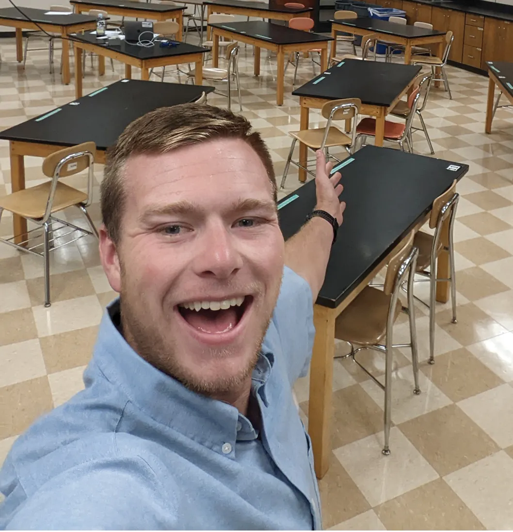 A man smiles while taking a selfie inside a classroom, gesturing toward rows of empty lab tables and chairs arranged neatly behind him.