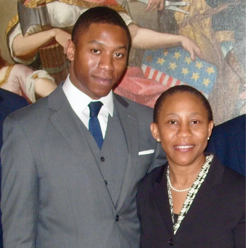 A formally dressed man and woman pose together indoors in front of a mural featuring historical imagery, smiling toward the camera during an official or ceremonial event.