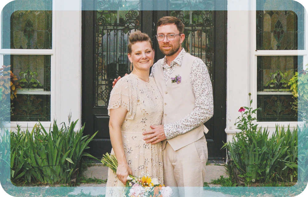 A smiling bride and groom pose together outdoors in wedding attire in front of a glass and wrought-iron door.