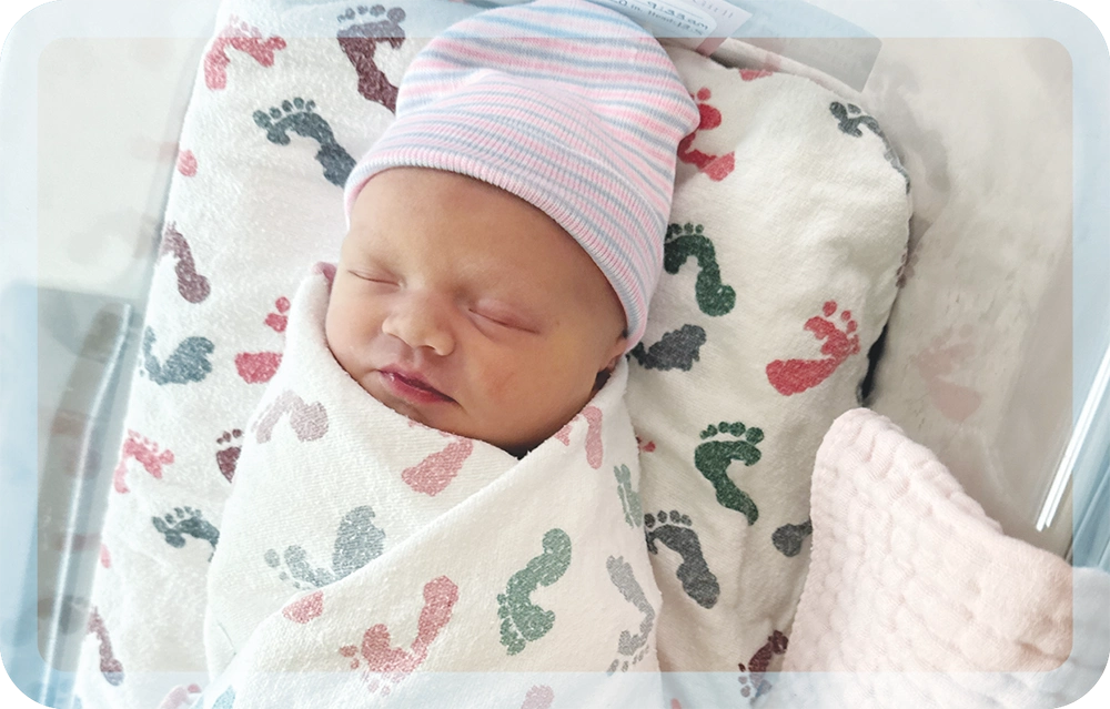 A newborn baby wearing a pink and blue striped hat sleeps swaddled in a blanket printed with colorful footprints.