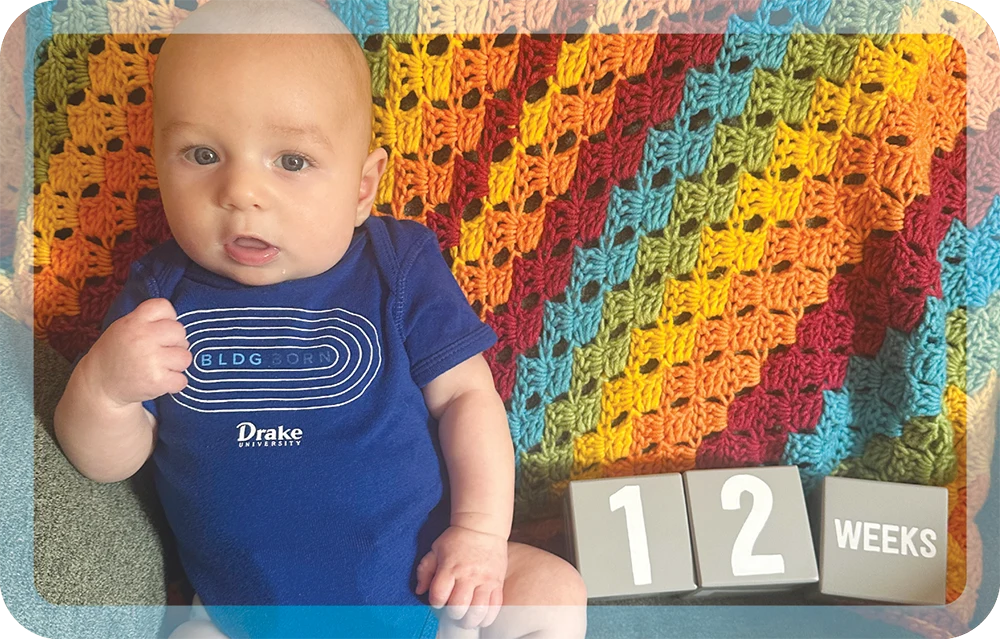 An infant in a blue Drake onesie lies on a rainbow blanket next to blocks reading "12 WEEKS."