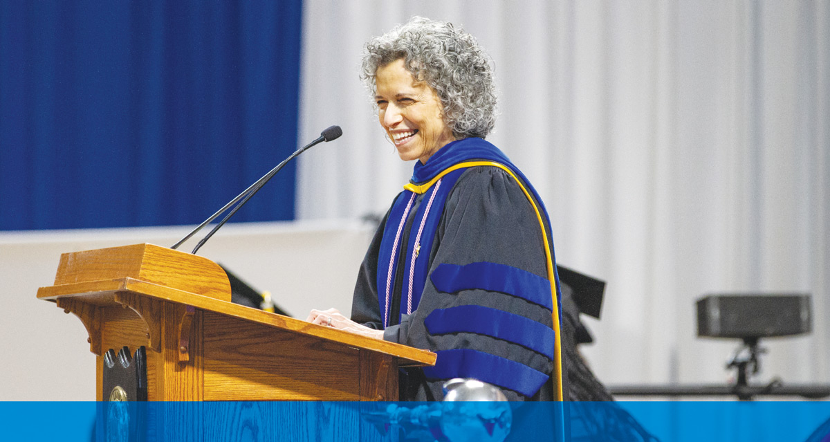 Sue Mattison in academic regalia speaks into microphones at a wooden podium.