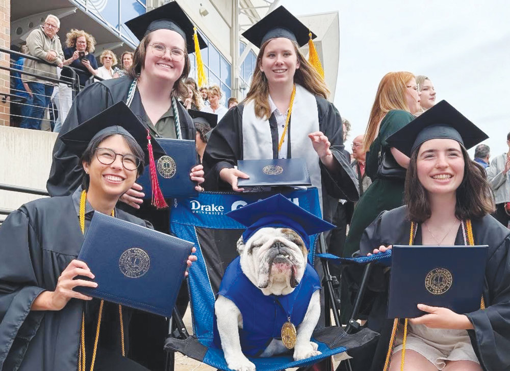 Four graduates in black caps and gowns pose with an English bulldog wearing a matching cap, gown, and medal.