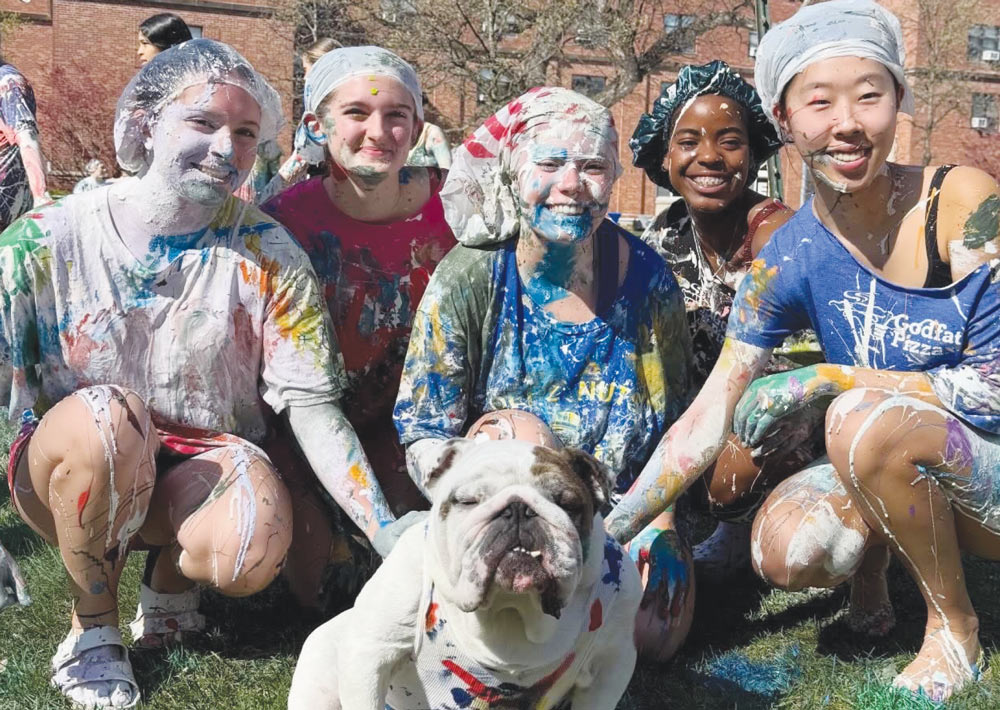 Five smiling students covered in colorful paint pose outdoors on the grass around an English bulldog.