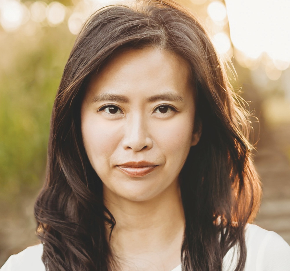 Close-up portrait of Ellen Won Steil with long dark hair wearing a white top, backlit by soft natural light outdoors.