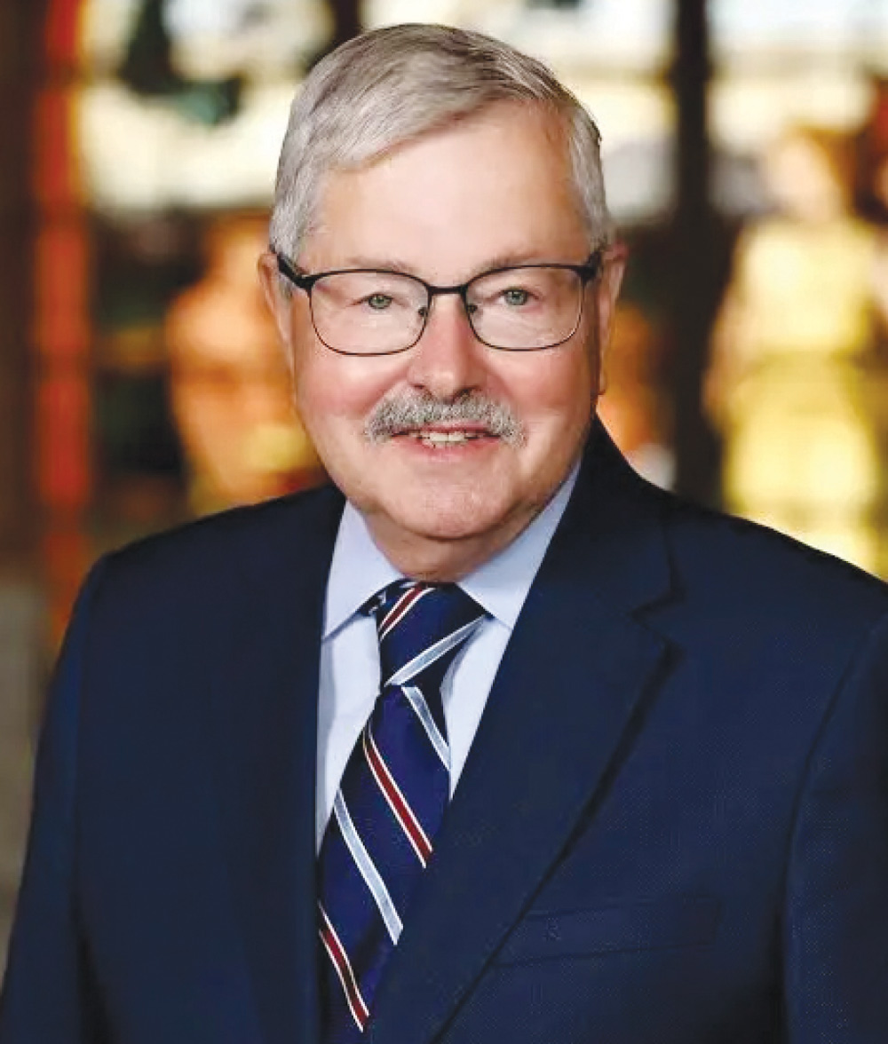 Terry E. Branstad wearing glasses and a mustache wears a blue suit jacket with a navy, red, and white striped tie.