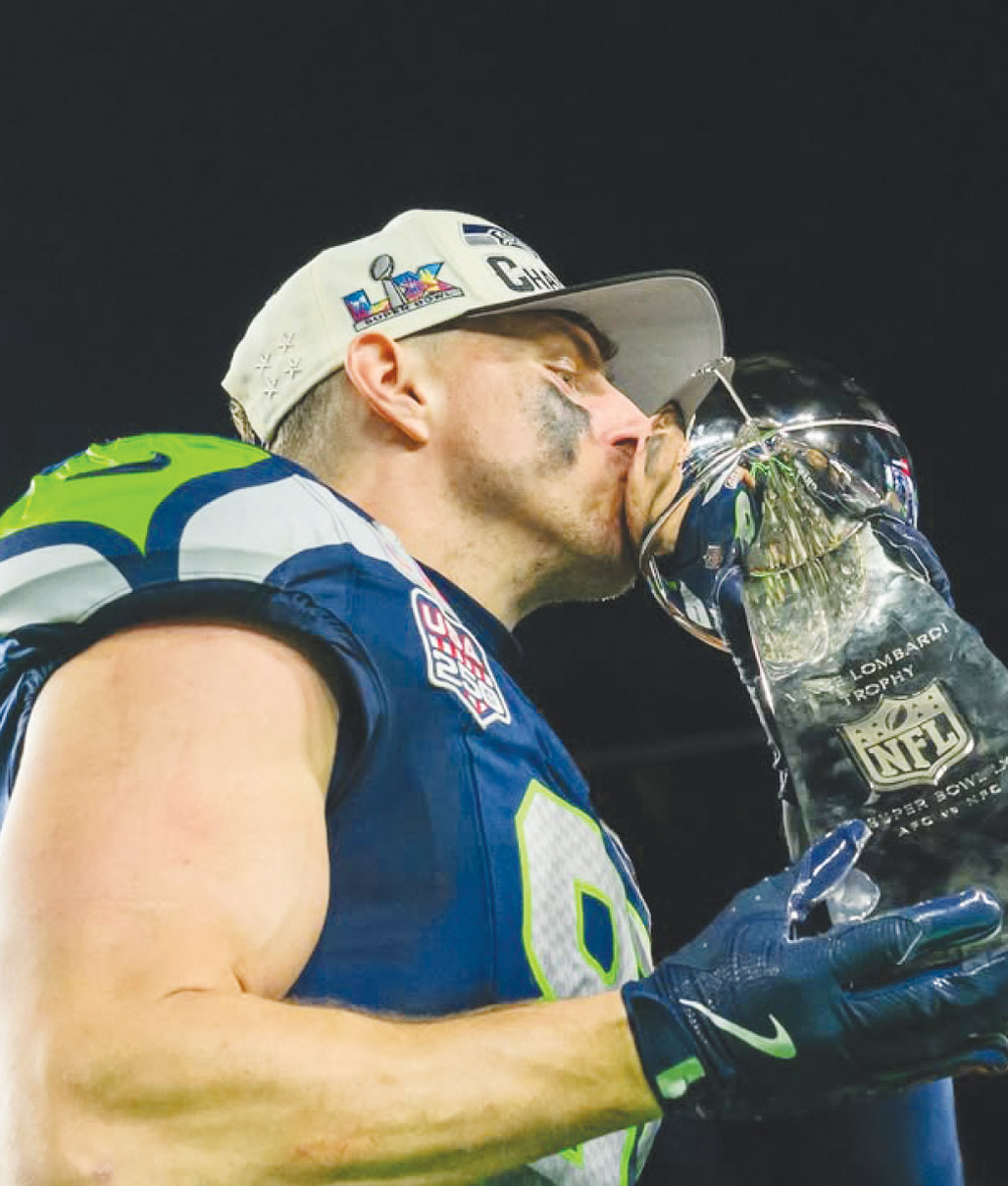 Eric M. Saubert wearing a Seattle Seahawks jersey and championship hat kisses the Vince Lombardi Trophy.