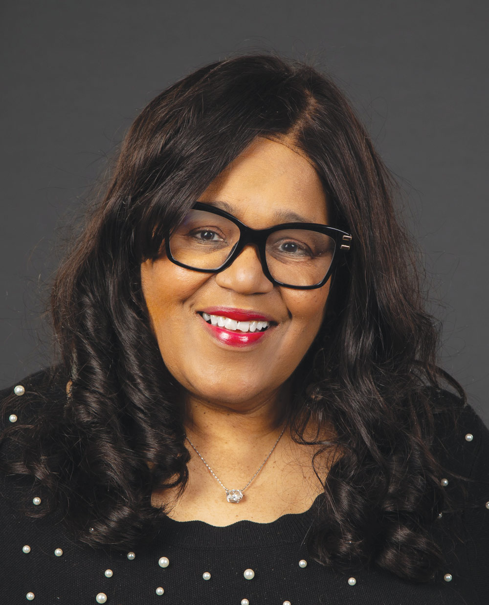Renee Hardman with dark curly hair and black glasses wears a black top decorated with white pearl accents.