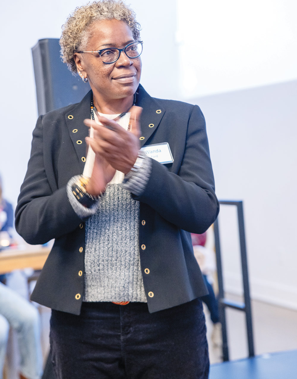 Wanda D. Ford with short gray hair and glasses claps while wearing a black blazer and grey sweater with a nametag.