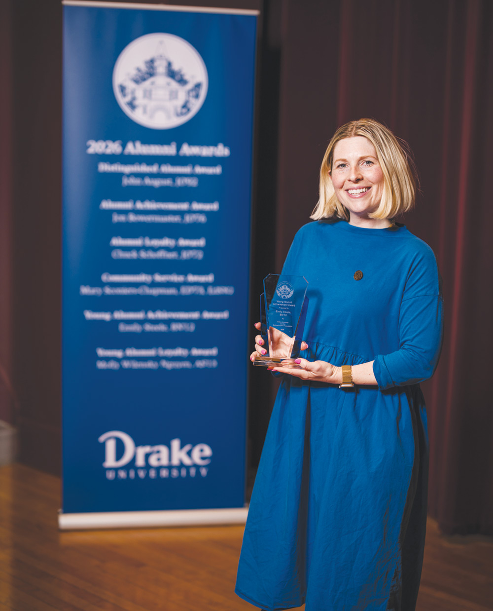 Emily A. (Boyd) Steele in a blue dress holds a clear award next to a Drake University Alumni Awards banner.