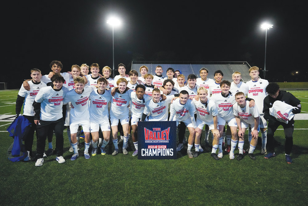 A men's soccer team in matching champion shirts poses on an outdoor turf field behind a conference sign.