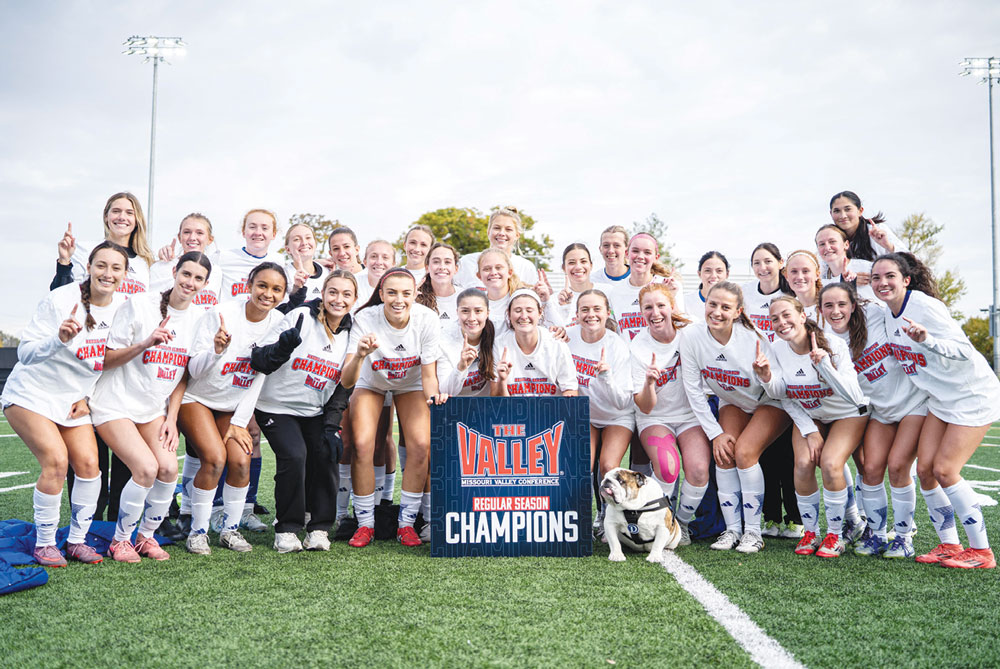 A women's soccer team in white champion shirts poses with their bulldog mascot on a field with a sign.