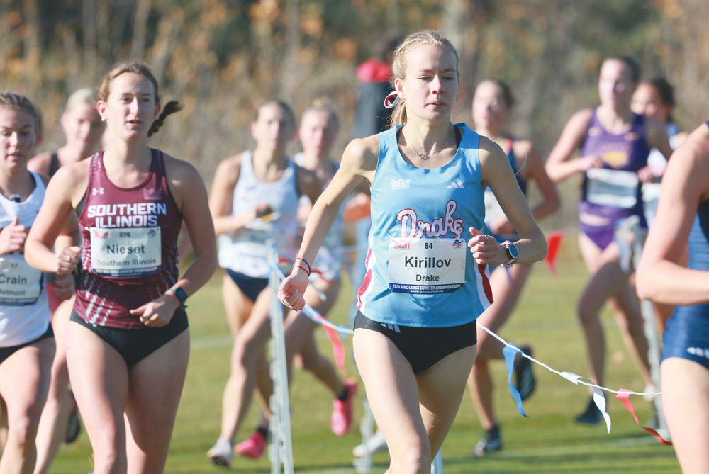 A female runner wearing a blue Drake bib and tank top competes in a cross-country race alongside opponents.