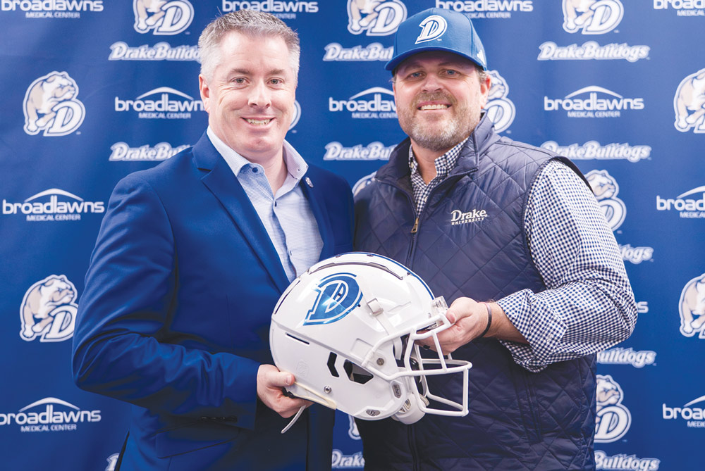 Two men in suits hold a white Drake Bulldogs football helmet together in front of a branded backdrop.