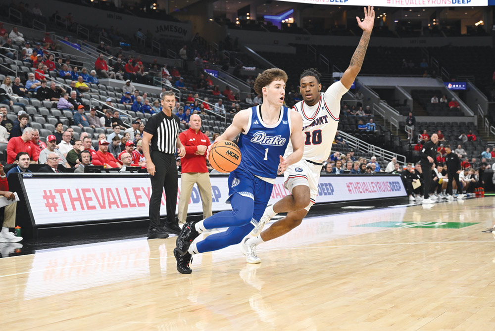 A basketball player in a blue Drake uniform drives past a defender during a game in a packed arena.