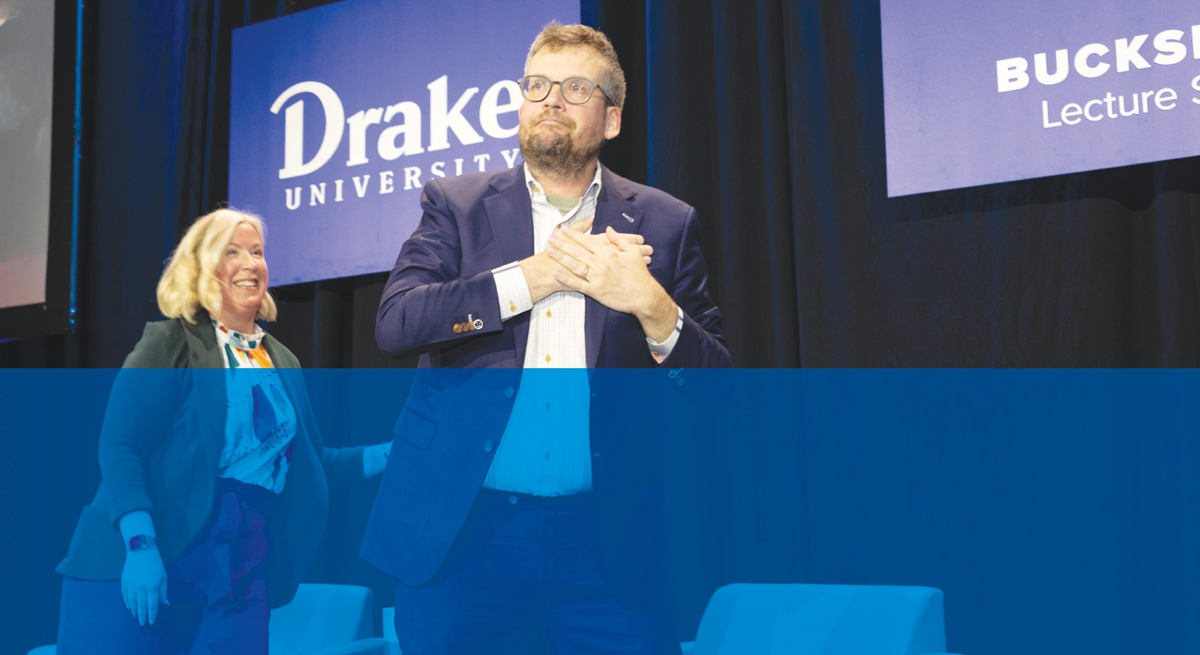 Author John Green holds his hands over his heart on stage next to a smiling woman in front of a Drake logo.