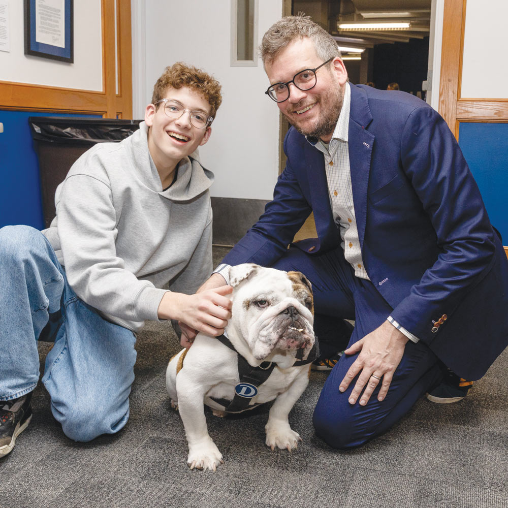 A young man and author John Green kneel on the floor together to pet an English bulldog.