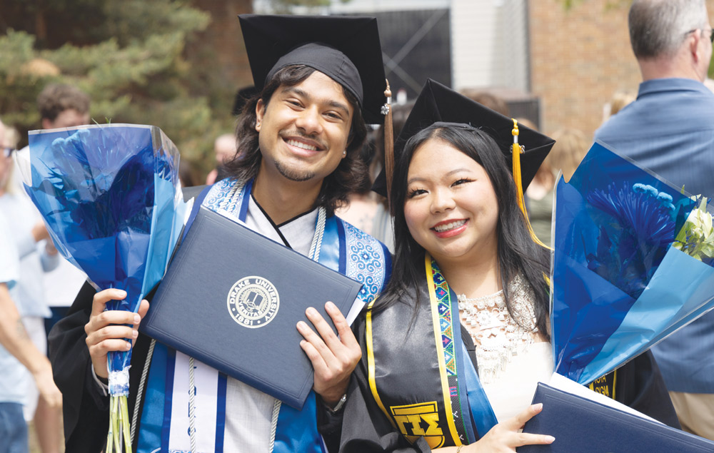Two smiling graduates in caps, gowns, and sashes hold blue flower bouquets and a Drake diploma cover.