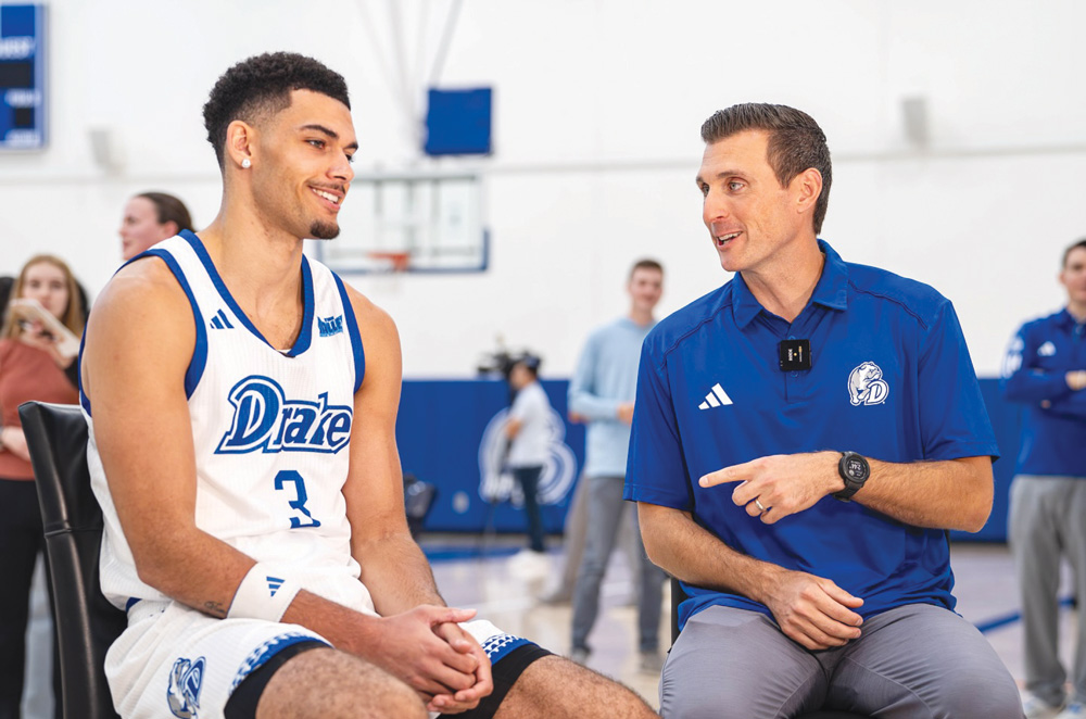 A Drake basketball player wearing number 3 sits and talks with a coach gesturing toward him during an interview.