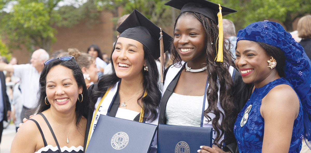 Four smiling graduates in black caps, gowns, and colorful headwear pose outdoors with diploma covers.
