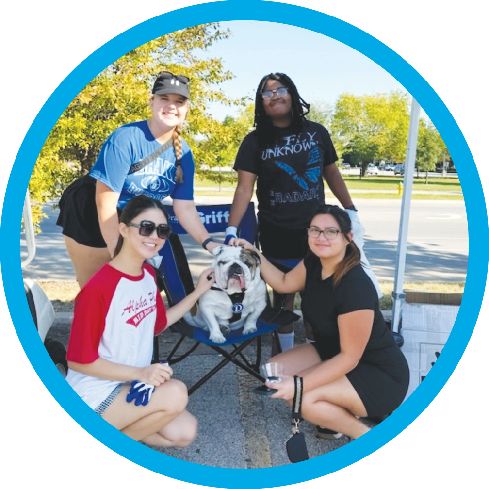 Four students pose around an English bulldog sitting in a chair outdoors inside a circular blue frame.