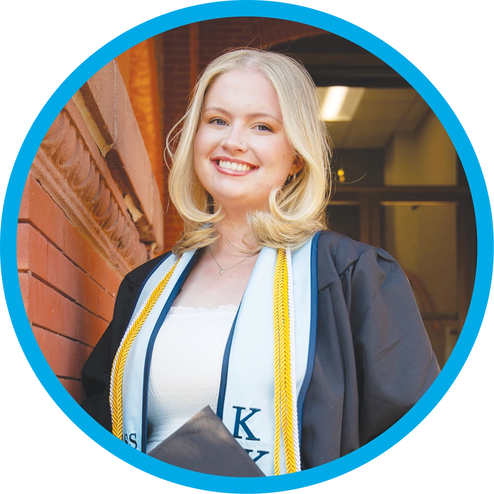 Emerson Whittemore wearing a cap, gown, and stoles smiles outside against a brick campus building.