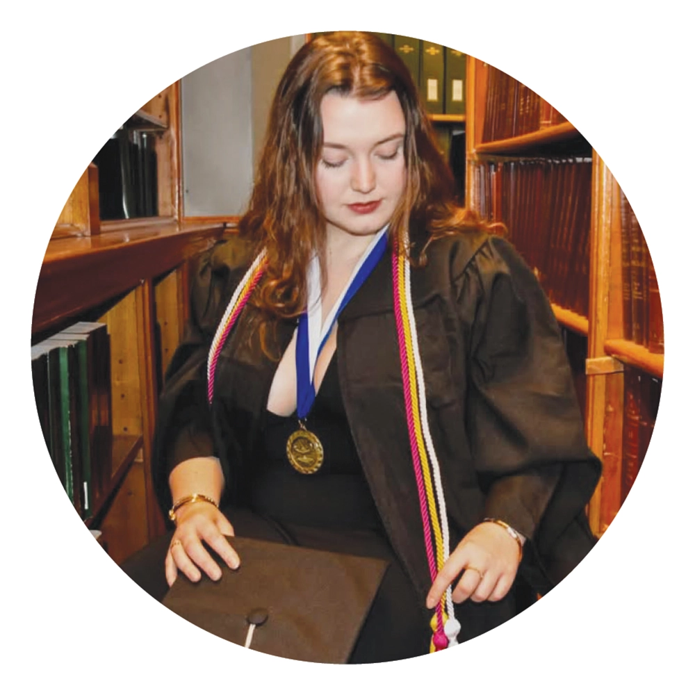 Keira O’Brien wearing a cap, gown, and honors cords sits in a library holding her mortarboard.