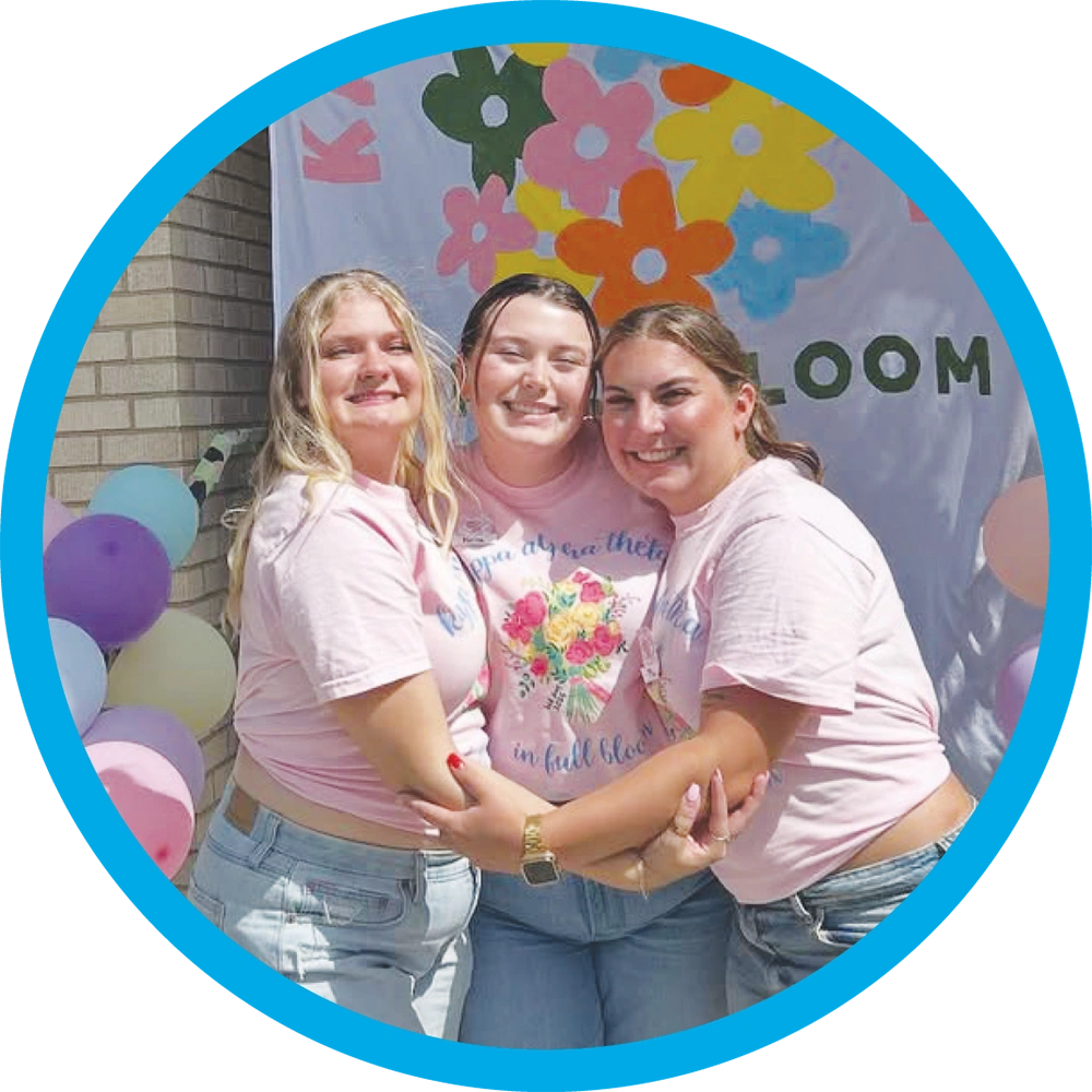 Three young women wearing matching pink shirts embrace and smile in front of a colorful banner.