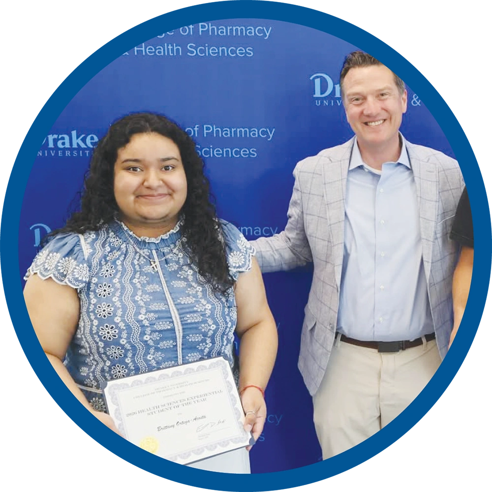 Brittany Ortega holds an award certificate alongside a smiling man in a blazer in front of a blue Drake backdrop.