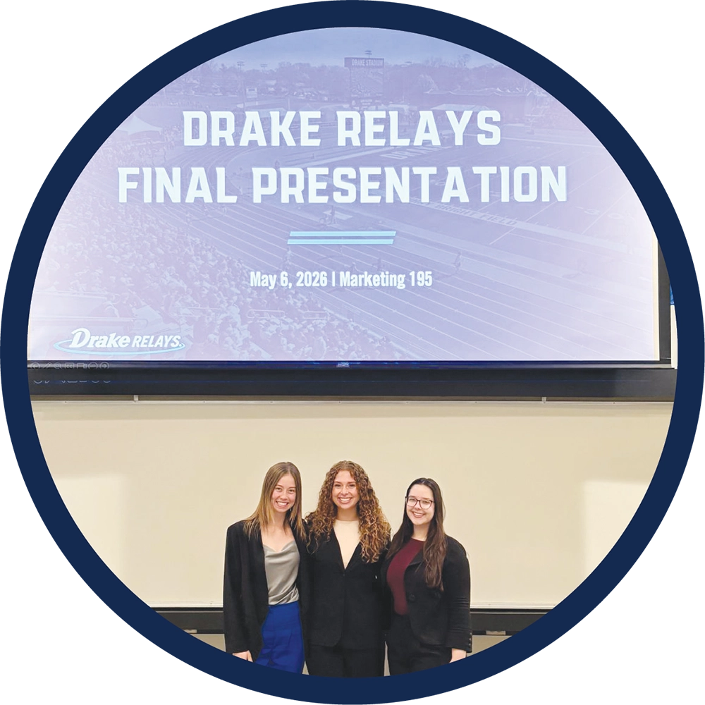 Three women in professional attire stand together in front of a presentation screen reading "Drake Relays".
