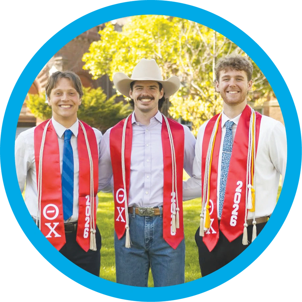 Three male students wear red graduation sashes outdoors, with the center student wearing a cowboy hat.
