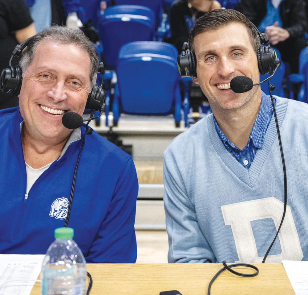 Two smiling sports broadcasters wearing headsets sitting at a commentary table inside a stadium arena.