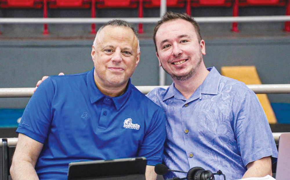 Two men smiling at a commentary table inside an arena, one wearing a blue Drake Bulldogs polo shirt.