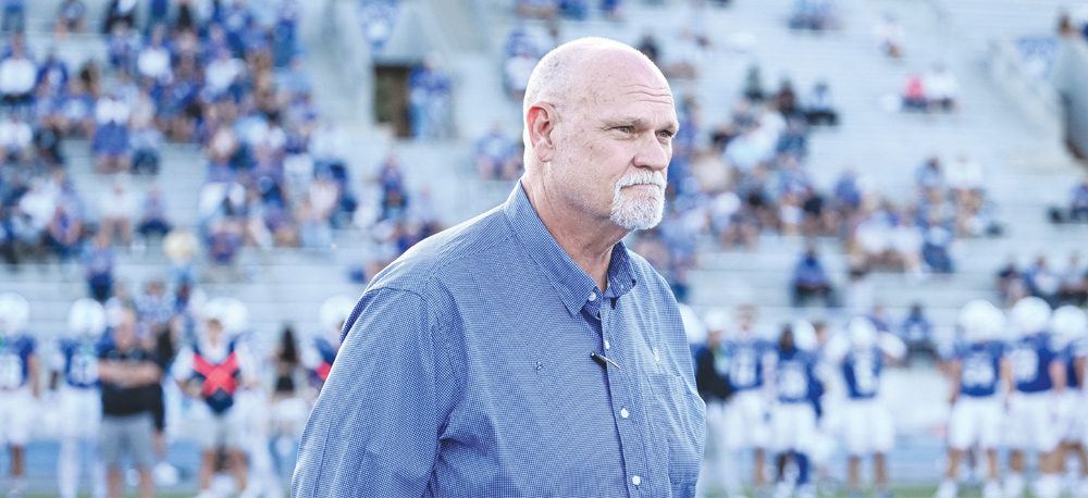 A man with a goatee in a blue button-down shirt stands outdoors on a football field with bleachers behind him.