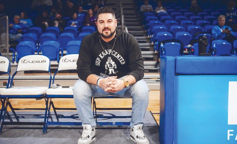 A man wearing a black hoodie and jeans sits on a court-side chair in an empty basketball arena.