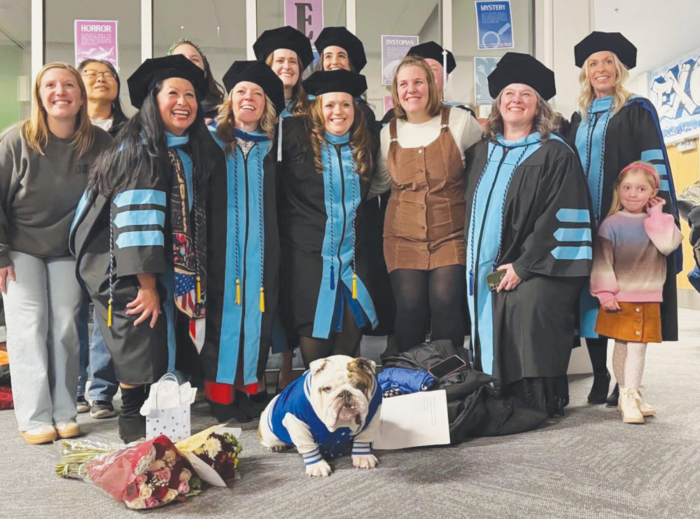 A group of graduates in academic regalia pose indoors around an English bulldog wearing a varsity jacket.