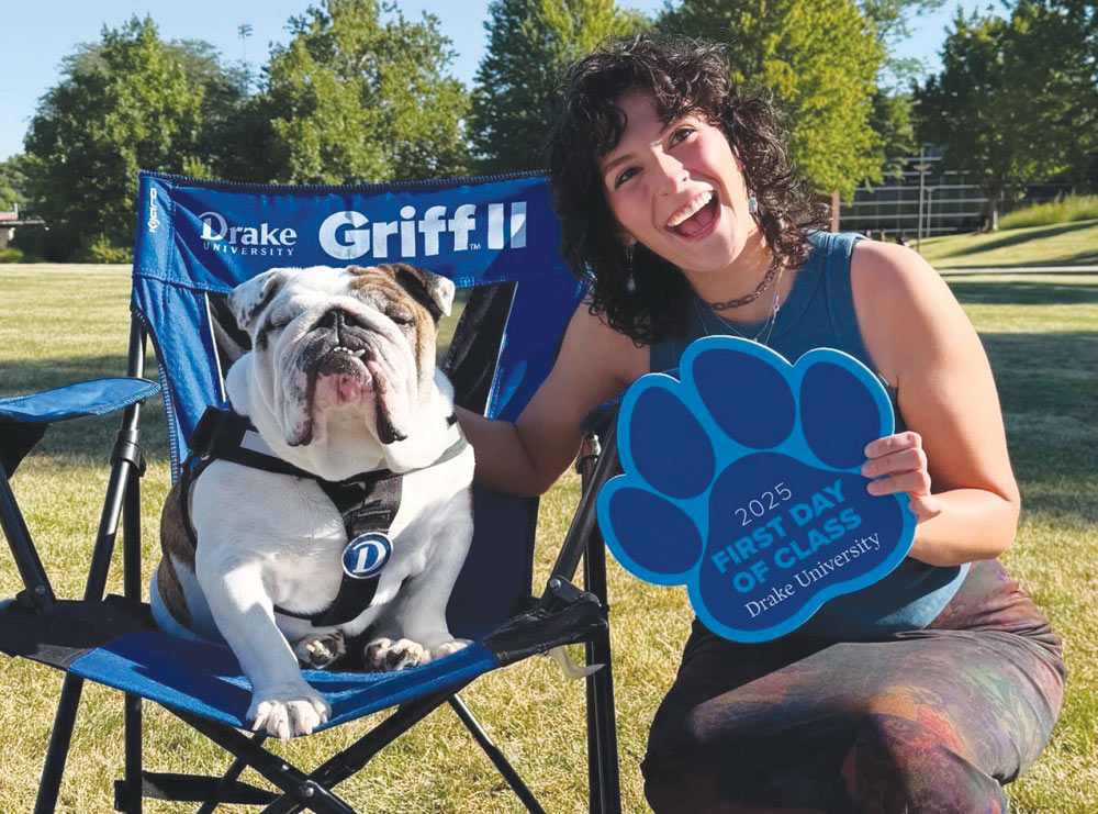 A smiling student holding a blue paw sign poses outdoors next to an English bulldog sitting in a chair.